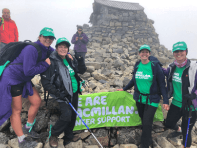 Four women in green Macmillan gear pose with a banner on a misty mountain summit.